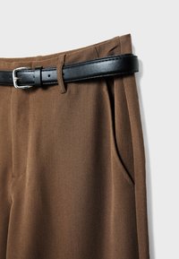 Pantalon marron avec détail de poche latérale et une ceinture en cuir noire passant dans les passants sur un fond clair.