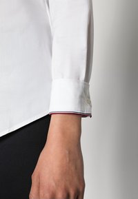 Left arm wearing white long sleeve shirt with buttoned cuff featuring red, white, and blue striped trim, against a neutral background.