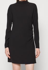 Femme portant une robe noire à manches longues avec un col haut et un léger froncé sur le côté, debout devant un fond uni.