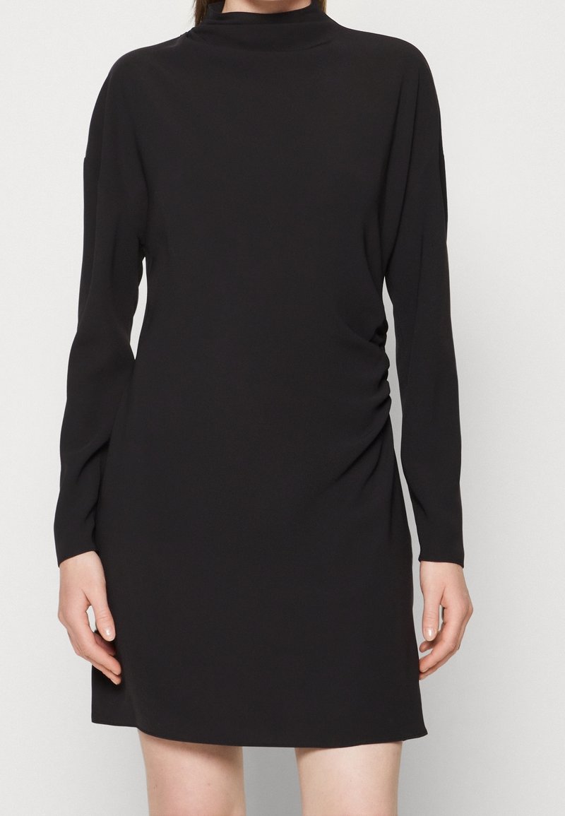 Femme portant une robe noire à manches longues avec un col haut et un léger froncé sur le côté, debout devant un fond uni.