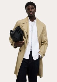 Beige Trenchcoat mit großen Revers und doppelreihiger Front, getragen über einem weißen Hemd und dunklen Jeans. Schwarze Tasche in der linken Hand gehalten.