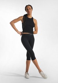 Sort ribbet crop top kombineret med sorte capri leggings. Modellen bærer hvide sportssko. En ensfarvet hvid baggrund fremhæver outfittets detaljer.