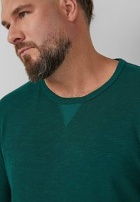 Donkergroen, gestructureerd shirt met een ronde hals en een driehoekige accent op de kraag. Korte mouwen, zacht materiaal.
