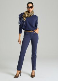 Vrouw in een donkerblauwe trui en broek, met een schuilprint sjaal en hakken, een bruine riem, en grote zonnebril, poserend tegen een effen achtergrond.