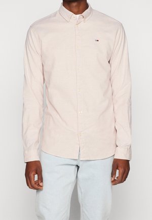 Homme portant une chemise beige clair à manches longues boutonnée avec un petit logo Tommy Hilfiger sur la poitrine et un jean bleu clair.