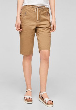 Person, die knielange beigetöne Shorts mit Kordelzug und weiße Plateausandalen trägt, steht vor einem einfachen Hintergrund.