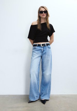 Ung kvinde iført sort off-shoulder top, lyseblå vidde bukser med sort bælte, sorte spidse sko og rektangulære solbriller, stående op ad en hvid væg.