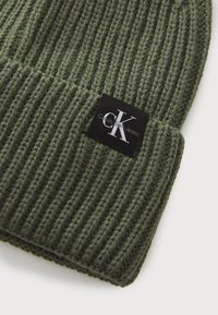 Olijfgroene gebreide beanie met ribbelstructuur en een omgeslagen zoom. Bevat een zwart etiket met witte branding aan één kant.