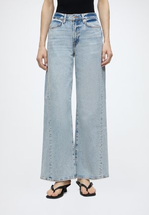 Person iført lyseblå højtaljede brede jeans, sort ærmeløs top og sorte sandaler, stående mod en ensfarvet baggrund.