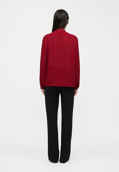 Femme aux longs cheveux foncés portant un chemisier rouge à manches longues et un pantalon noir, se tenant de dos sur un fond clair uni.