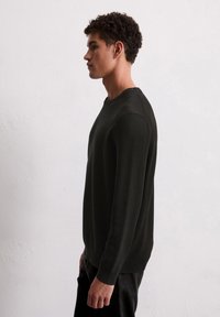 Jeune homme aux cheveux bouclés portant un pull noir ajusté à manches longues et un pantalon noir, se tenant de profil contre un mur blanc.