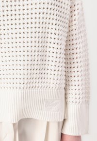 Maglione bianco lavorato a maglia con motivo a trama aperta, orlo a costine e dettagli testurizzati. Presenta una patch con il logo sul lato inferiore.
