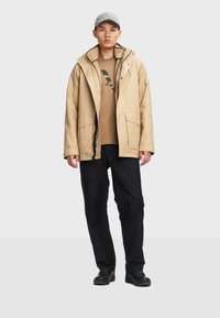 Beige wasserdichte Jacke mit flauschigem Innenfutter, kombiniert mit einem braunen graphic T-Shirt und schwarzen Hosen, ergänzt durch eine graue Mütze und schwarze Schuhe.