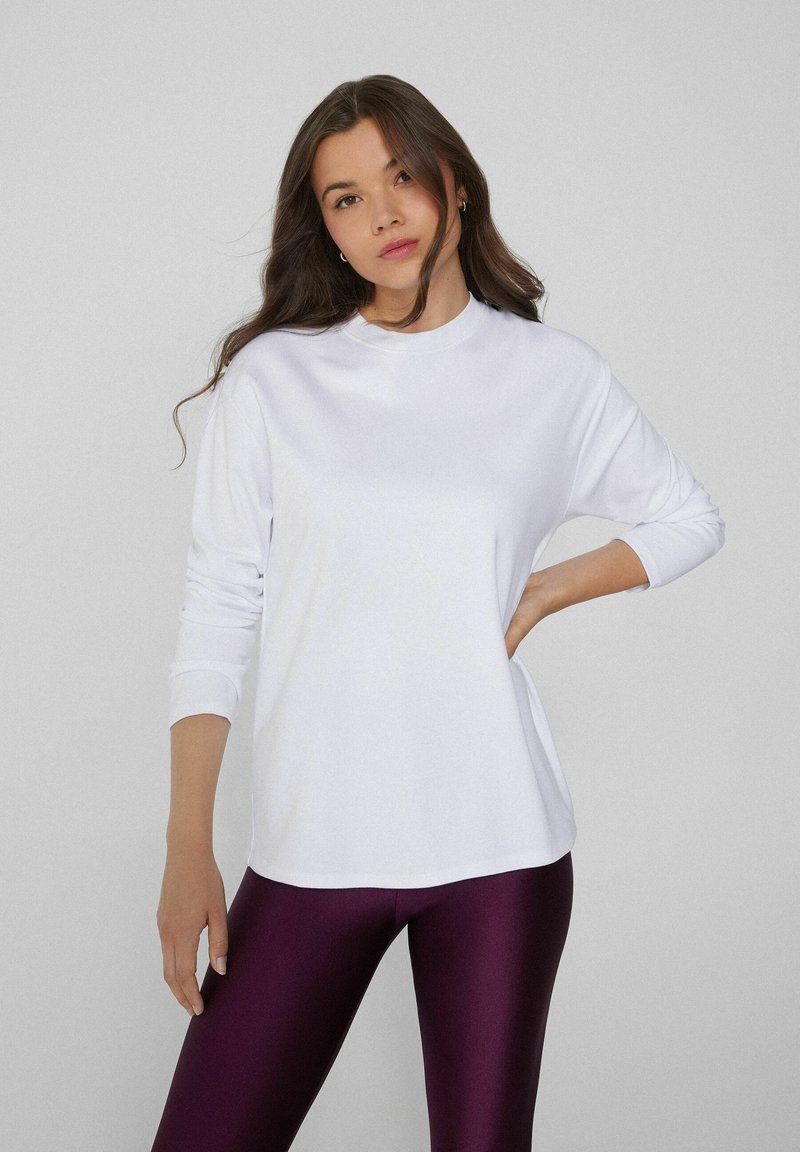Weißes Langarmoberteil mit Rundhalsausschnitt und lässiger Passform, kombiniert mit glänzenden lila Leggings; glatte Textur und schlichtes Design.