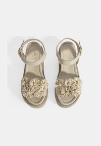 Beige espadrille sandalen met een platte zool, voorzien van een bloemige versiering en enkelbandje. Het materiaal lijkt zacht met een textuur.