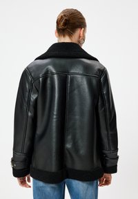 Kvinna med håret i en knut bär en svart läderjacka med fårskinnskrag och manschetter, i kombination med blå jeans, visad bakifrån mot vit bakgrund.