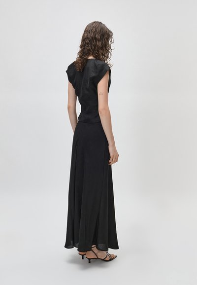 Γυναίκα με σγουρά μαλλιά φοράει μαύρο αμάνικο τοπ, μακριά μαύρη φούστα και μαύρα σανδάλια με τακούνι, στέκεται πλάτη σε απλό φόντο.