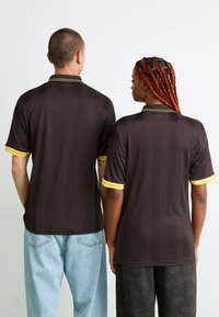 Hnedé polokošele s krátkym rukávom a textúrovaným vzorom, s žltými akcentmi na golieri a rukávoch, nosené dvoma osobami zozadu.