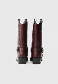 Bordo krāsas ādas kaubojzābaki ar gravētiem ziedu rakstiem, sānu siksnām un ķēdes akcentiem. Stiprās blokpapēži, velkama dizains bez šņorēm.