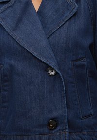 Κοντινό πλάνο σκουρόμπλε τζιν μπουφάν με γιακά πέτου, δύο μαύρα κουμπιά και μπροστινή τσέπη με ορατές ραφές.