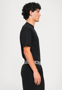 Νέος ά�νδρας με σγουρά μαύρα μαλλιά, φορώντας μαύρη μπλούζα και μαύρο παντελόνι με λευκή και μαύρη ζώνη Versace Jeans Couture, στραμμένος προς τα δεξιά.