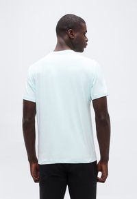 Άνδρας που κοιτάζει προς τα πίσω, φοράει κοντομάνικη ανοιχτό μπλε μπλούζα και μαύρο παντελόνι, με απλό ανοιχτό φόντο.