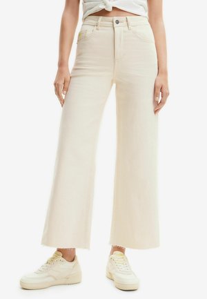 Personne portant un jean crème à jambes larges avec ourlets bruts, des baskets blanches et un haut blanc noué à la taille, debout devant un fond uni.