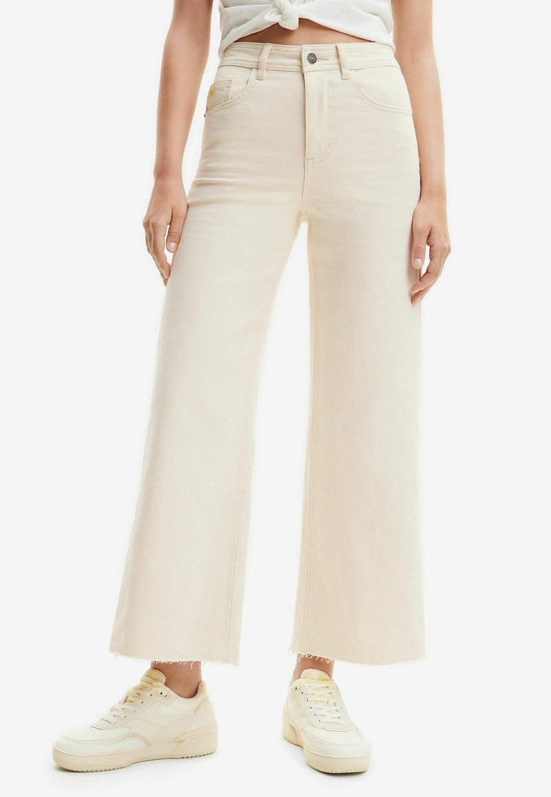 Personne portant un jean crème à jambes larges avec ourlets bruts, des baskets blanches et un haut blanc noué à la taille, debout devant un fond uni.