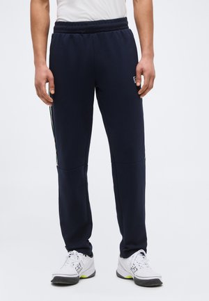 Άνδρας που φοράει ναυτικού μπλε φόρμες με ελαστική μέση και άσπρα αθλητικά παπούτσια, στέκεται μπροστά σε λευκό φόντο.