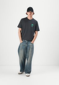 T-shirt en coton gris foncé avec un logo vert sur la poitrine, associée à un jean bleu clair à coupe ample et une casquette noire, complétée par des chaussures blanches.