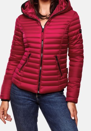 Kobieta ubrana w dopasowaną, czerwoną pikowaną kurtkę z kapturem i kieszeniami na zamek, w zestawie z niebieskimi dżinsami.