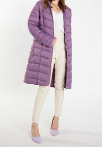 Kvinde iført en lang lilla dunjakke, en hvid top med blonder, cremefarvede bukser og lavendel høje hæle på en hvid baggrund.