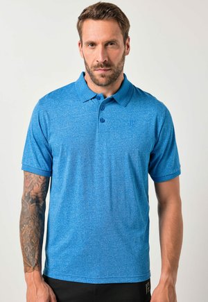 Mand med kort hår og skæg iført en blå polo-shirt og sorte bukser, stående med venstre arm med tatoveringer synlig mod en ensfarvet baggrund.