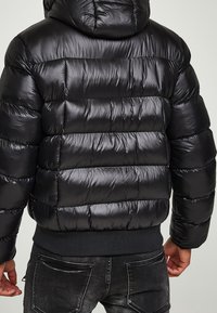 Zwarte pufferjas met een glanzende, gewatteerde buitenkant, voorzien van een capuchon en een geribbelde zoom. Het ontwerp bevat opvallende horizontale stiknaden.