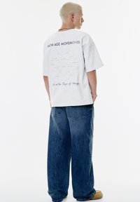 Grå oversized t-shirt med et tekstureret geometrisk mønster på ryggen og trykt tekst. Stylet med løse blå denim jeans.
