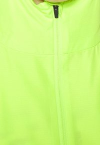 Veste néon verte brillante en matériau léger, dotée d'une fermeture éclair centrale et d'un col montant. Texture lisse sans motifs visibles.