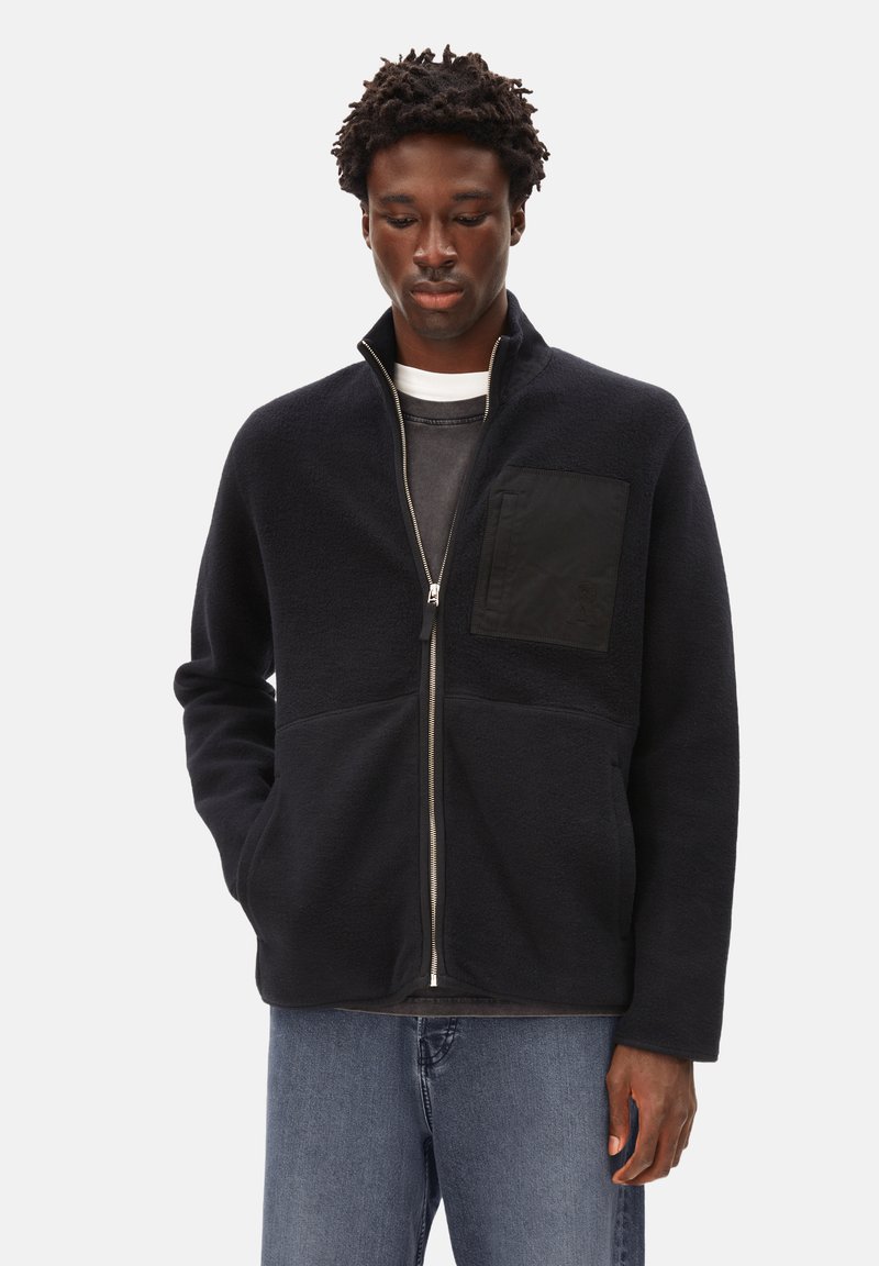 Marineblaue Fleecejacke mit hohem Kragen, Frontreißverschluss und einer schwarzen Stoff-Putztasche auf der Brust. Strukturierte Oberfläche, lässige Passform.