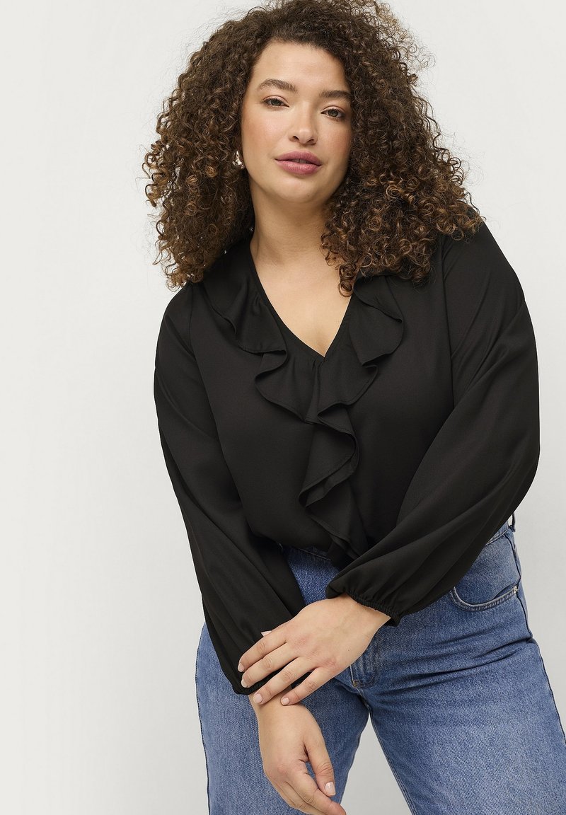 Schwarze Bluse mit V-Ausschnitt und Rüschendetails, langen, puffigen Ärmeln, kombiniert mit blauen Denim-Jeans. Glatte Stofftextur.