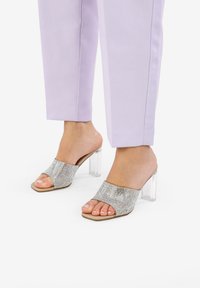Sandalias de punta abierta adornadas con brillantes plateados y un tacón bloque transparente, combinadas con pantalones ajustados de color púrpura pastel.