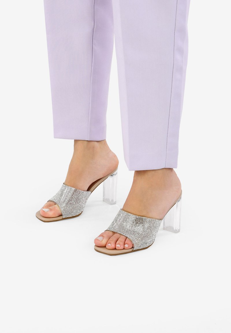 Sandalias de punta abierta adornadas con brillantes plateados y un tacón bloque transparente, combinadas con pantalones ajustados de color púrpura pastel.