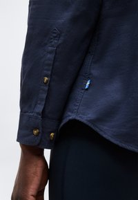 Sötétkék hosszú ujjú ing mandzsettája barna gombokkal és egy kis, varrott svéd zászlós címkével a személy karján, a oldalsó varrás közelében.