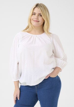 White, textured blouse with gathered neckline and three-quarter sleeves; styled with blue jeans, featuring a relaxed fit and subtle dotted pattern.