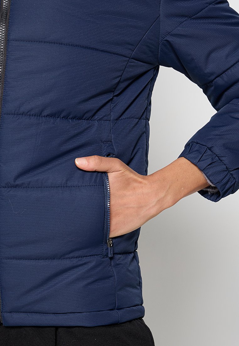 Mano inserita nella tasca laterale con zip di un giubbotto trapuntato blu navy con polsini elastici sulle maniche.