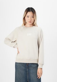 Sudadera de color beige claro con mangas raglán, puños y dobladillo acanalados, que presenta el logo de "GAP" en blanco en el pecho. Textura simple y suave.