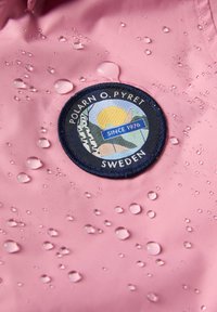 Vedenkestävä vaaleanpunainen kangas, jossa on pyöreä kangasmerkki vuorten, auringon ja tekstin "POLARN O. PYRET" sekä vuoden "1976" kuvastamalla. Vedetippoja näkyy.