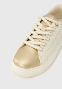 Hvid sneaker med metallisk guld tåhætte, teksturerede snørebånd og logodetalje på siden. Glat materiale med lavt design.