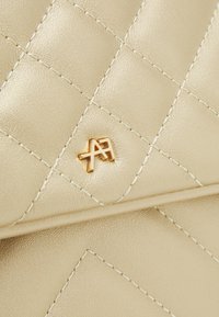 Beige quiltat läder med logotypdetalj i guldfärg, med vita sömmar och en slät yta. Närbild.