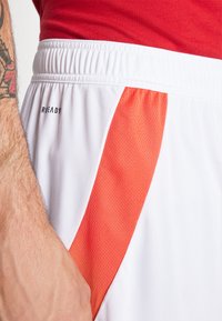 Baltos sportinės šortai turi tekstūruotą oranžinį intarpą, elastinę liemenį ir atspausdintą logotipą. Artimas vaizdas paryškina medžiagą ir pasiuvimą.