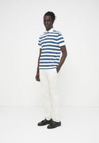 Ριγέ πόλο πουκάμισο με μπλε, λευκές και ναυτικές ρίγες, με γιακά και μικρό λογότυπο. Συνδυάζεται με λευκό παντελόνι και μαύρα παπούτσια.