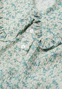 Kukallinen paita vaaleansinisessä ja vihreässä, valkoisilla napilla ja frilla-kauluksella. Pehmeä, teksturoitu kangas, jossa on pieniä apiinoita.